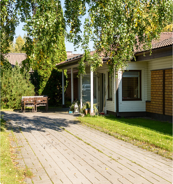 Päällystetty ajotie johtaa yksikerroksiseen taloon, jossa on valkoiset sivuraiteet ja tiiliaksentit ja jota ympäröivät vihreät puut ja ruoho. Puinen penkki istuu vasemmalla pensaiden lähellä varjossa.