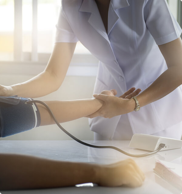 Valkoiseen univormuun pukeutunut terveydenhuollon ammattilainen tarkistaa potilaan verenpaineen mansetin ja monitorin avulla klinikalla, kun taustalla ikkunasta paistaa auringonvalo.